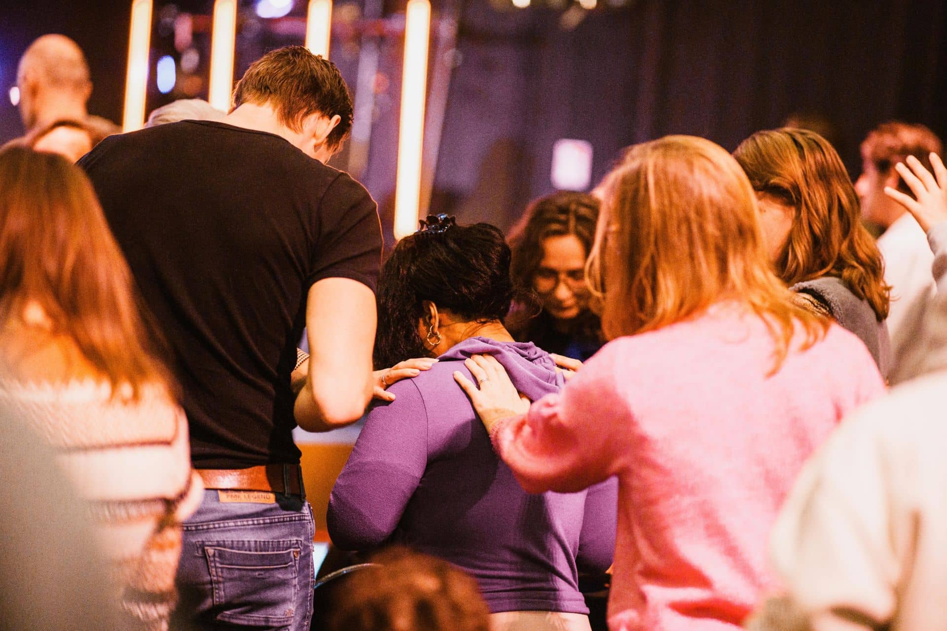 A group of people gathers closely, with several individuals placing their hands on another person's shoulders in a supportive gesture.
